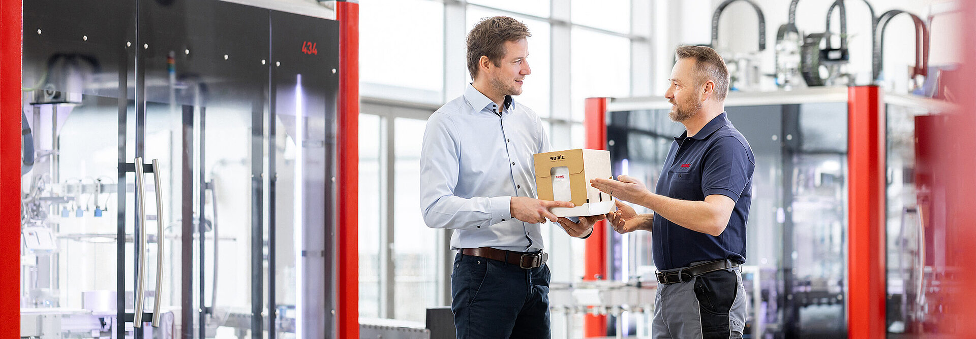 Zwei Mitarbeiter begutachten einen Kartonzuschnitt vor einer SOMIC Karton-Verpackungsmaschine in der Inbetriebnahmehalle – Verpackungsberatung von SOMIC